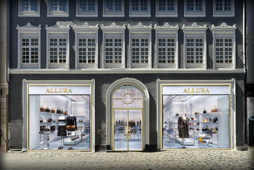 Building displays luxury clothing and bags in storefront windows labeled "ALLURA," situated on an urban street.