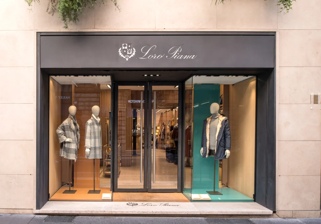Mannequins display coats behind glass storefront of clothing store labeled "Loro Piana," set in urban environment.