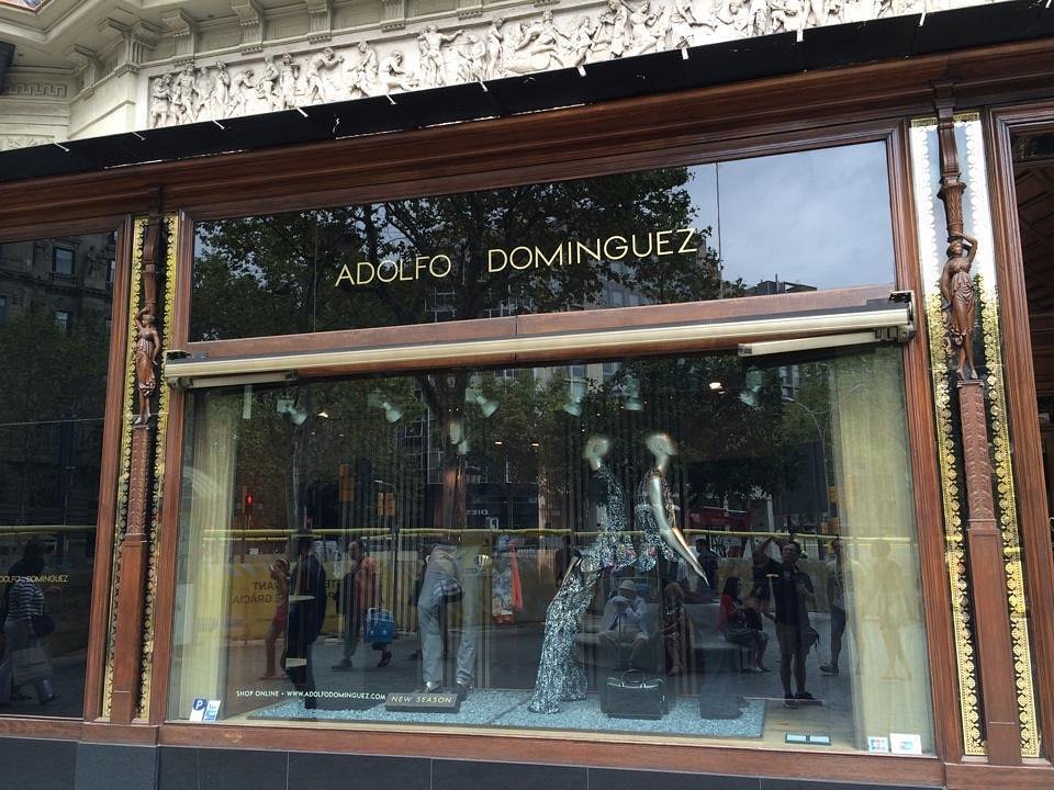 Mannequin displaying clothes, sits in a store window of Adolfo Dominguez, with reflections of people on the street visible.