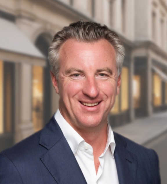 A person smiling, wearing a suit, in front of a blurred urban background. Ian Kelley