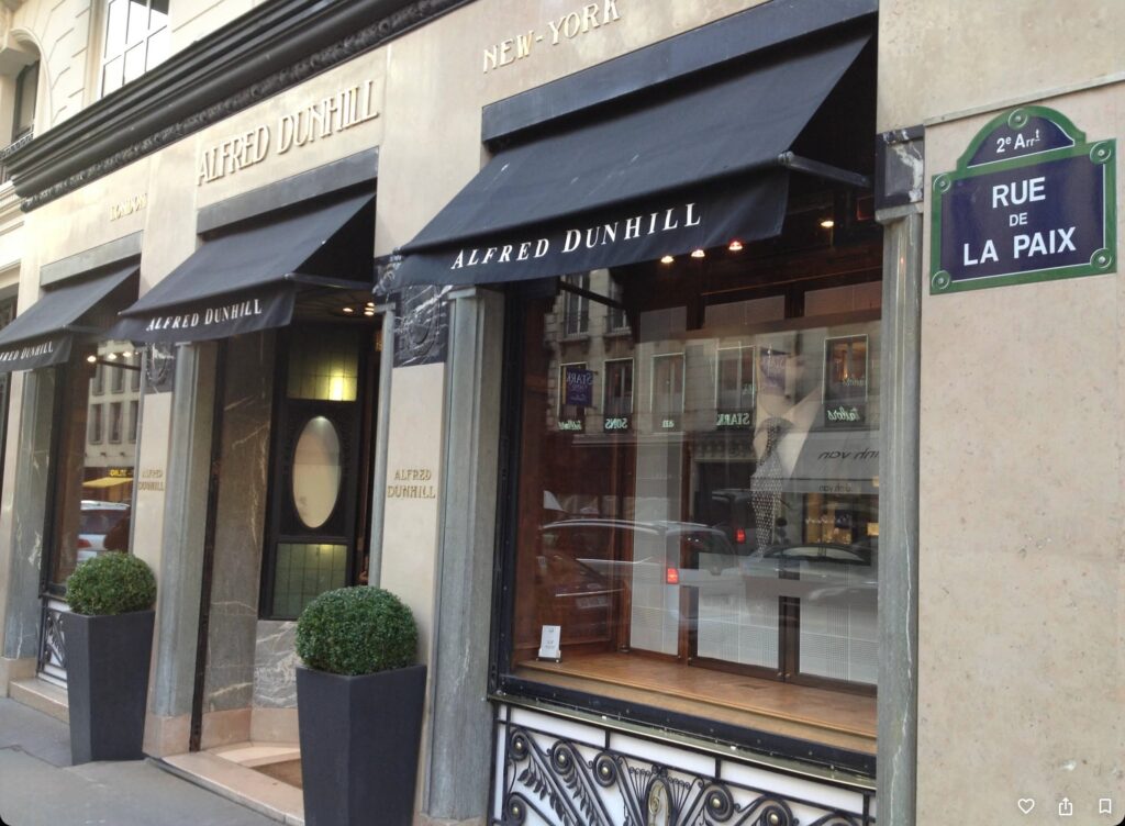 Storefront displays "Alfred Dunhill" signage, with reflective windows and potted plants, located on Rue de la Paix in an urban environment.