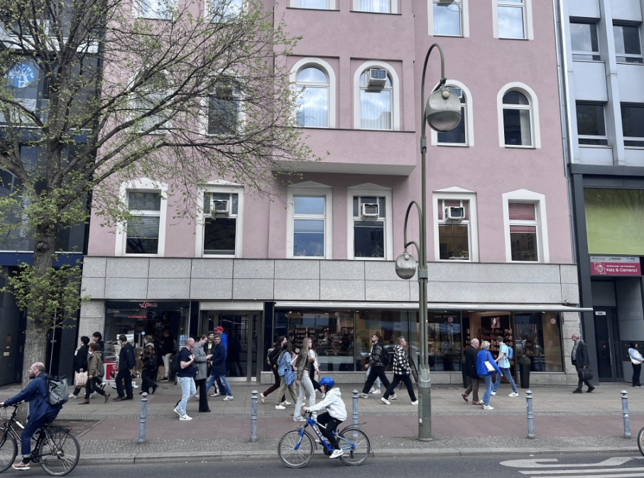 People walking and cycling on sidewalk near a pink building with shops, trees, and street lamps. Text: "Altnatura Super Natur Markt," "Fass & Camera 1."