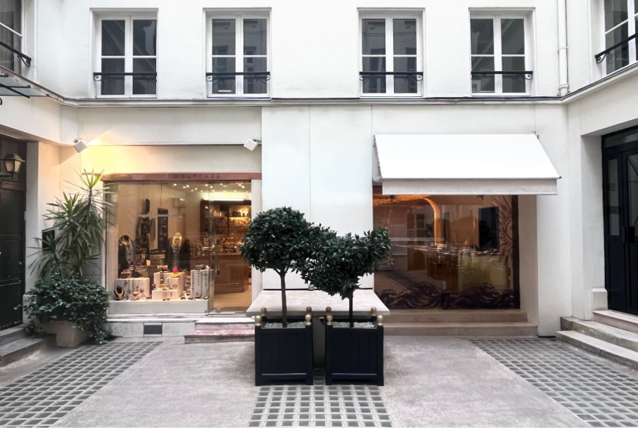Plants in planters stand in a courtyard, flanked by shopfronts displaying jewelry and decorated interiors.