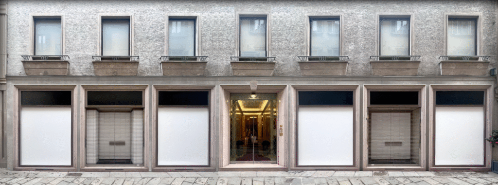 Building facade displaying large windows covered with white blinds, a central glass door, and six upper windows on a cobblestone street.