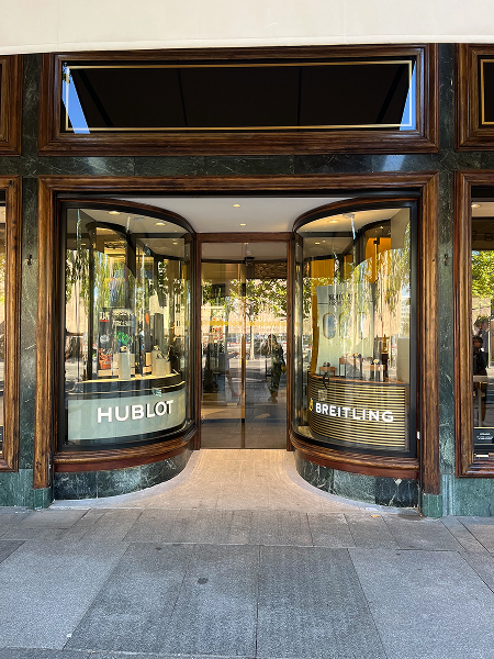 Storefront displays watches; curved windows showcase "HUBLOT" and "BREITLING"; situated in a city shopping area.